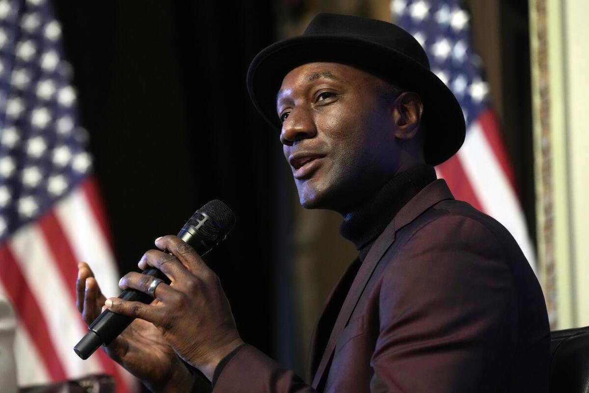 Aloe Blacc in a suit and a black hat speaks into a hand microphone in front of two American flags